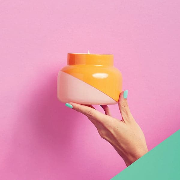 A hand is holding a squatty, round glass jar in front of a bright pink wall. The bottom corner of the background is a turquoise color. The model’s fingernails are painted the same turquoise color. The jar is split diagonally with the top half being a bright orange color and the bottom half being a soft pink color. The top of the jar is a lot narrower than the rest of the jar. There is a small flame inside the candle. 