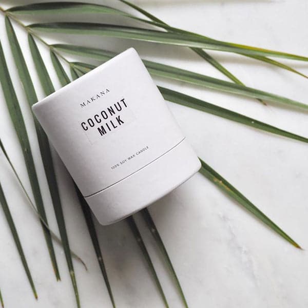 A clear glass jar candle with white wax and a white square label that reads, "Coconut Milk Makana" in clean black text. Photographed here is the outer packaging that features a white cylindrical cardboard package and features the same black text.