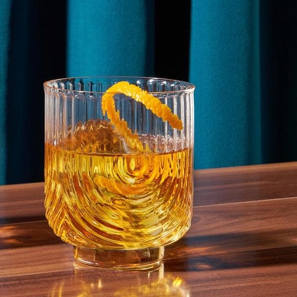 A clear glass tumbler with a gold base and an etched upside down arched design photographed here with a brown liquid and an orange peel garnish.