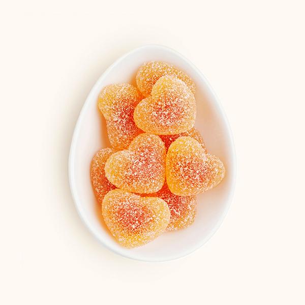 In front of a white background is a white egg shaped dish. The dish is filled with peach colored heart shaped gummies coated in white sugar. 