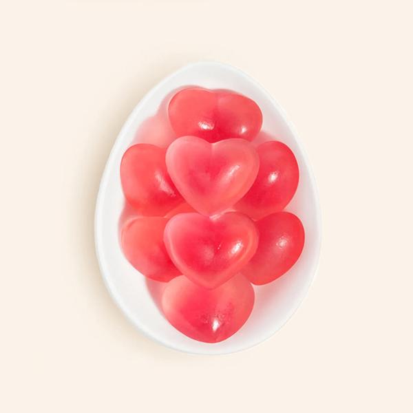In front of a peachy background is the birds eye view of a white egg shaped dish. The dish is filled with bright pink heart shaped gummies. 