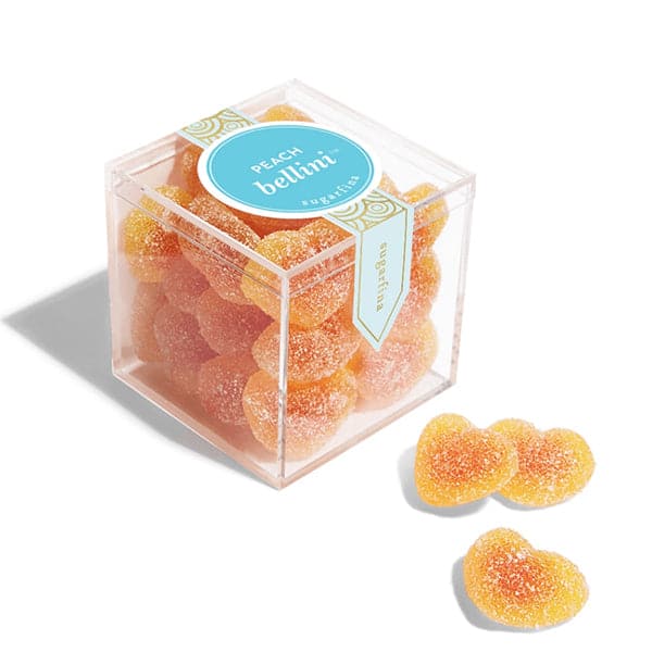 In front of a white background is a clear plastic cube container. The container is filled with peach colored heart shaped gummies coated in white sugar. On the top of the container is a bright blue circle sticker with white text that reads ‘peach Bellini.’ There are three gummies sitting outside of the container.