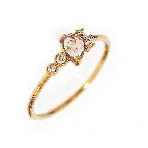 Against a white background is a slim, gold ring. On the front of the ring is a light pink crystal tear drop. Lining the right side of the tear drop are three white, round crystals. To the left of the tear drop is two white crystals with a gold border.