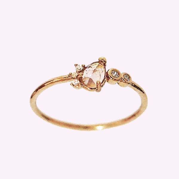 Against a soft pink background is a slim, gold ring. On the front of the ring is a light pink crystal tear drop. Lining the left side of the tear drop are three white, round crystals. To the right of the tear drop is two white crystals with a gold border.