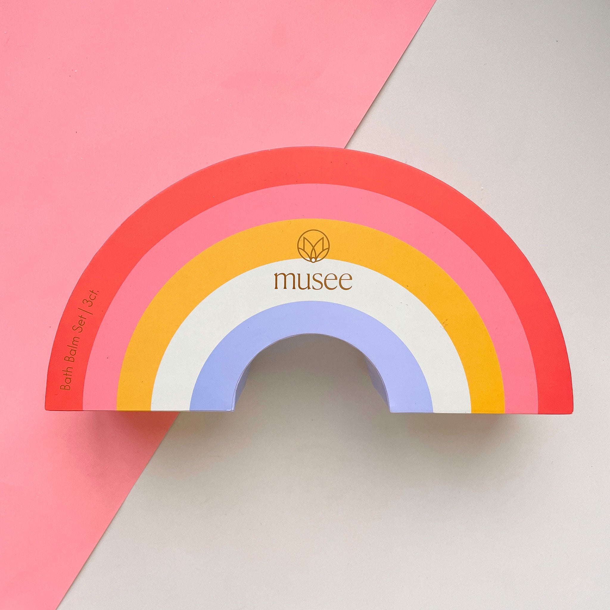 A set of bath bombs packaged inside of a rainbow box with pastel colors. The three bath bombs inside are a light shade of blue, pink and green.