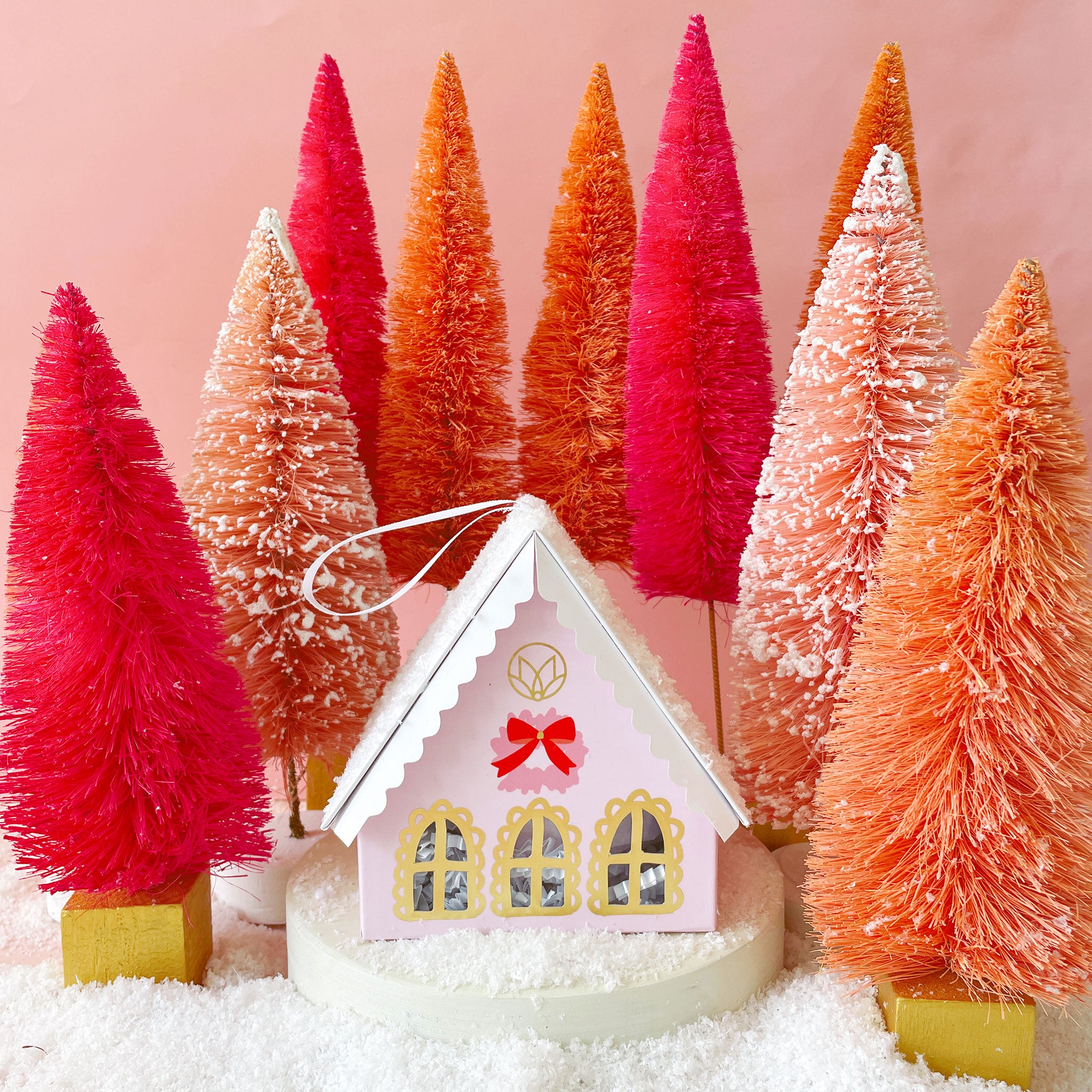 A pink holiday cottage that houses a pink bath bomb. The holiday cottage also doubles as an ornament with its ribbon loop for hanging. Bath bomb house surrounded by pink and orange tinsel trees on snow.