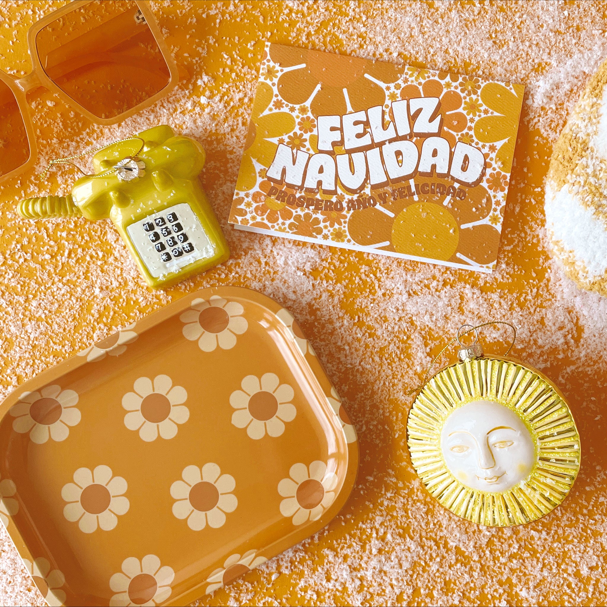 Greeting card filled with yellow and orange retro flower print. The card reads &#39;Feliz Navidad&#39; in white curved bubble letters. Below reads &#39;prosper año y felicidad&#39; in rust colored lettering. The card is accompanied by a solid white envelope. Card is surrounded by a glass sun ornament, glass yellow/green vintage phone ornament, a flower tray and a pair of Kelso sunglasses.