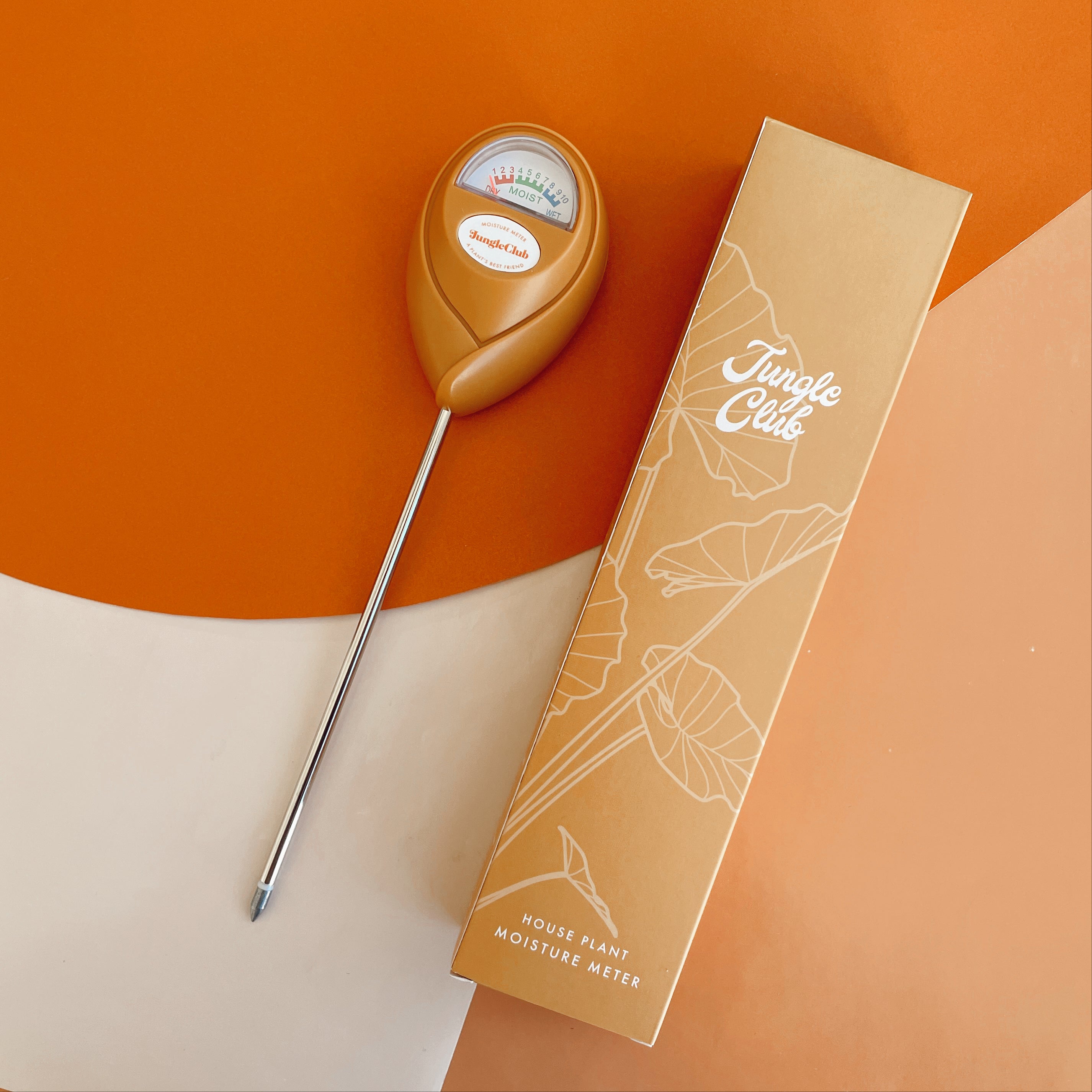 An orange moisture meter with dial at top that indicates dry moist of wet conditions for a plant lays on top of a bright orange background paper.  A peach colored paper intersects the right side of the image and the box that the moisture meter comes in sits on top of the paper. It reads "Jungle Club" and has the outlines of an alocasia grapically designed in white.