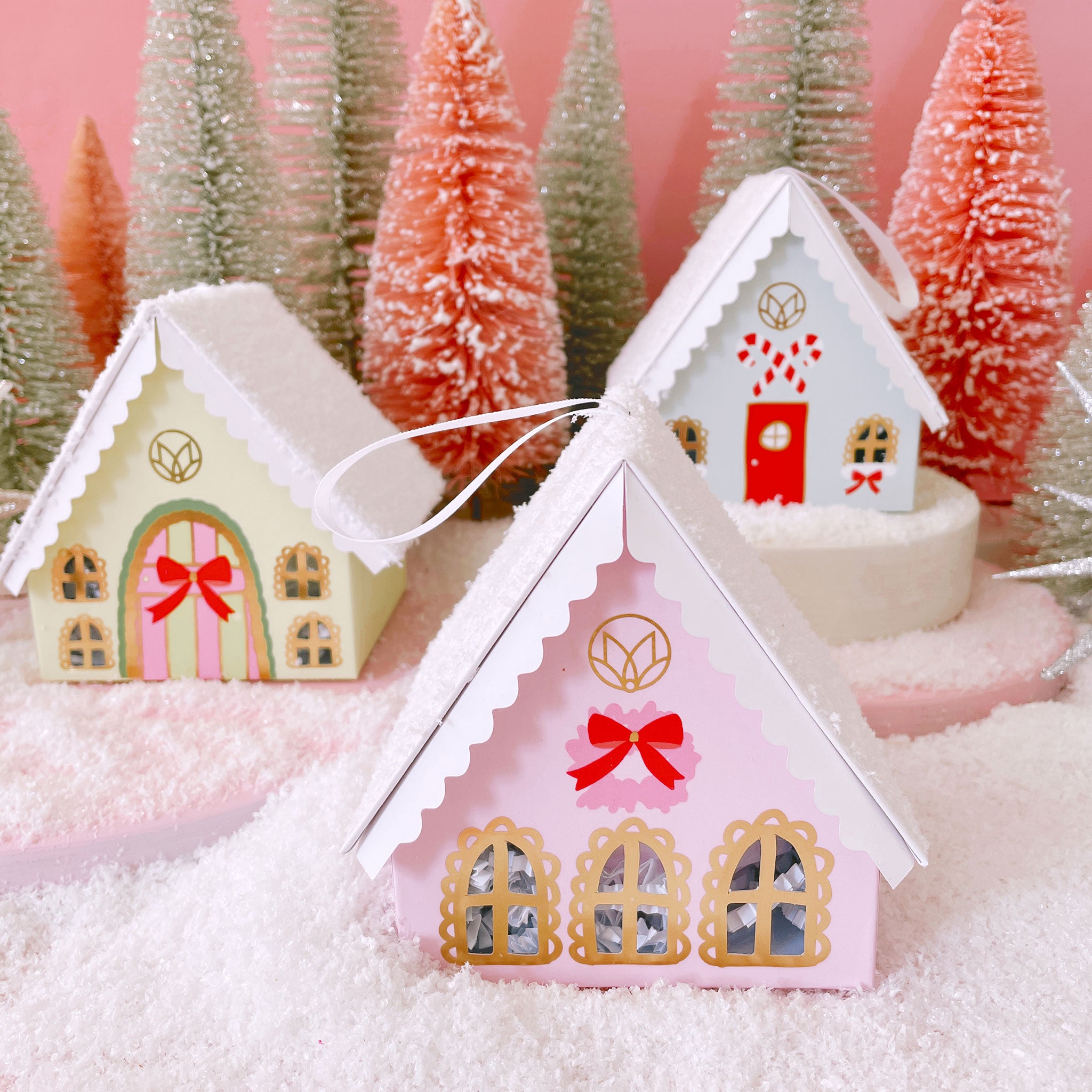 Green, pink and blue holiday cottages that is home to a blue bath bomb. The cardboard cottage comes with a ribbon loop at the top that makes it double as an ornament. Surrounded by silver and pink tinsel trees on snow.