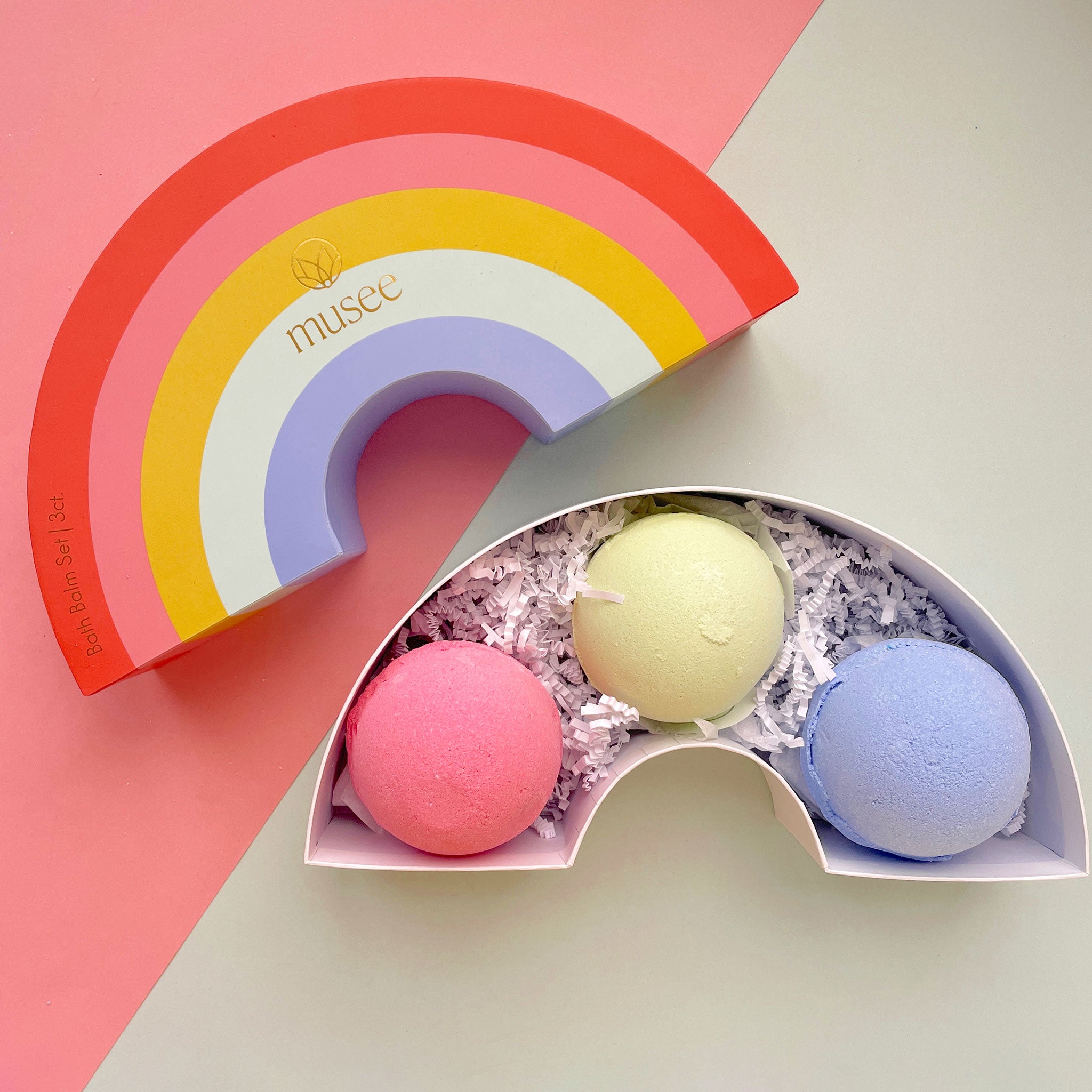 A set of bath bombs packaged inside of a rainbow box with pastel colors. The three bath bombs inside are a light shade of blue, pink and green.