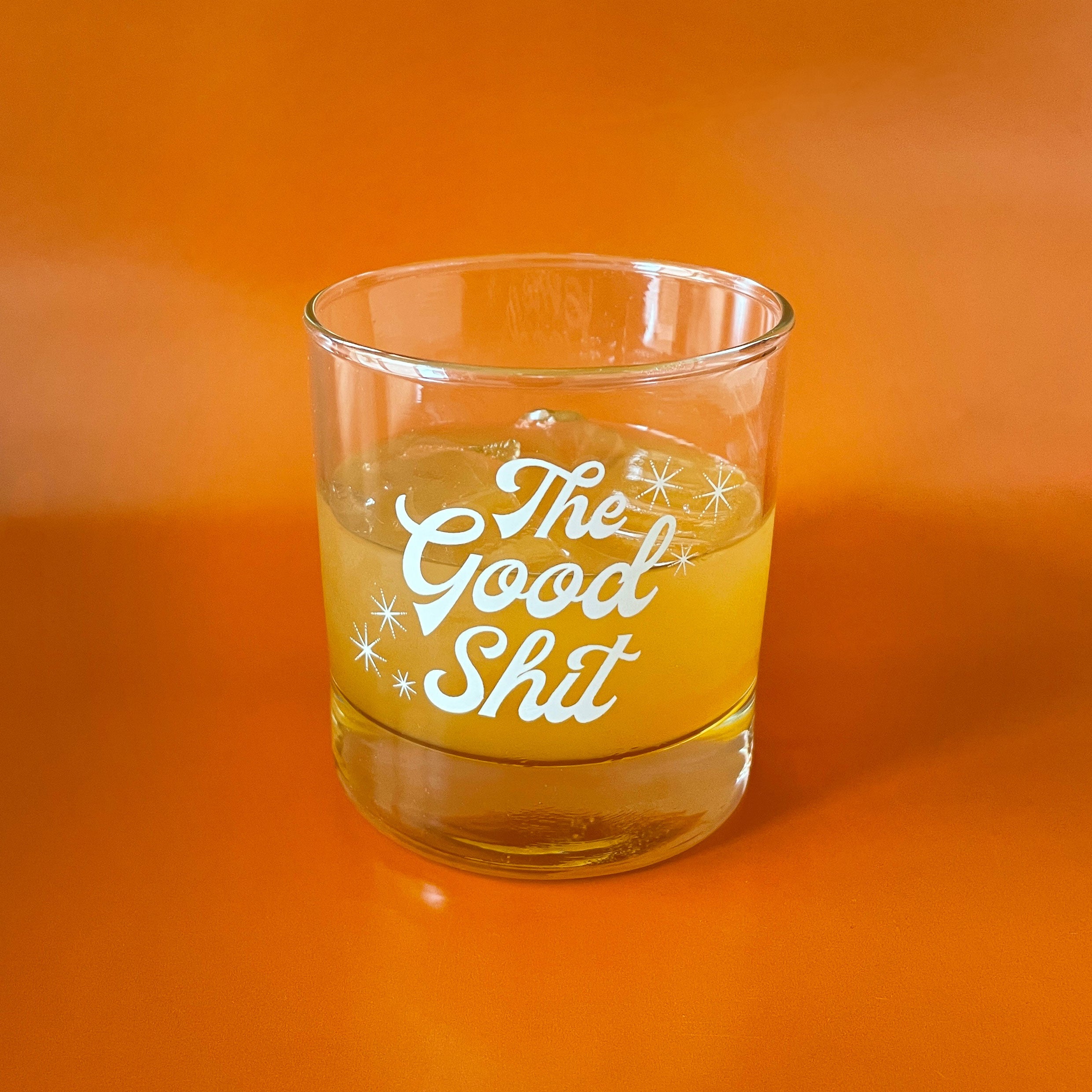 Against an orange background is a photograph of a short glass tumbler with a thick bottom and &quot;The Good Shit&quot; printed across the center in white groovy cursive text.