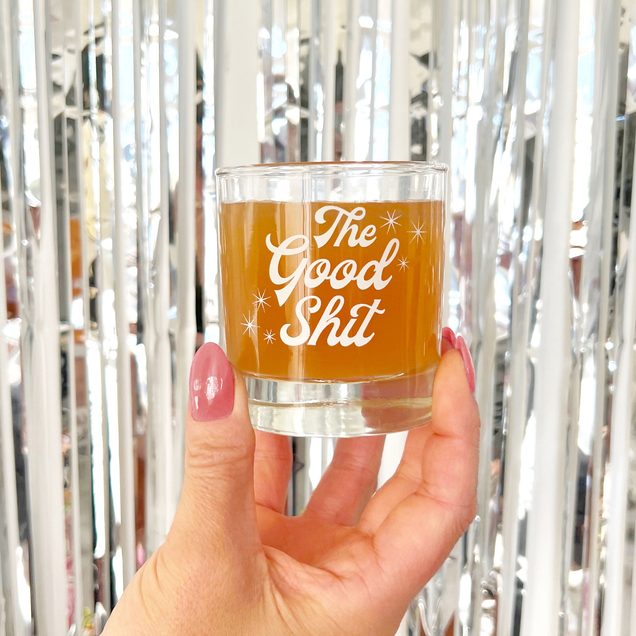Against a silver tinsel backdrop is a photograph of a short glass tumbler with a thick bottom and "The Good Shit" printed across the center in white groovy cursive text.