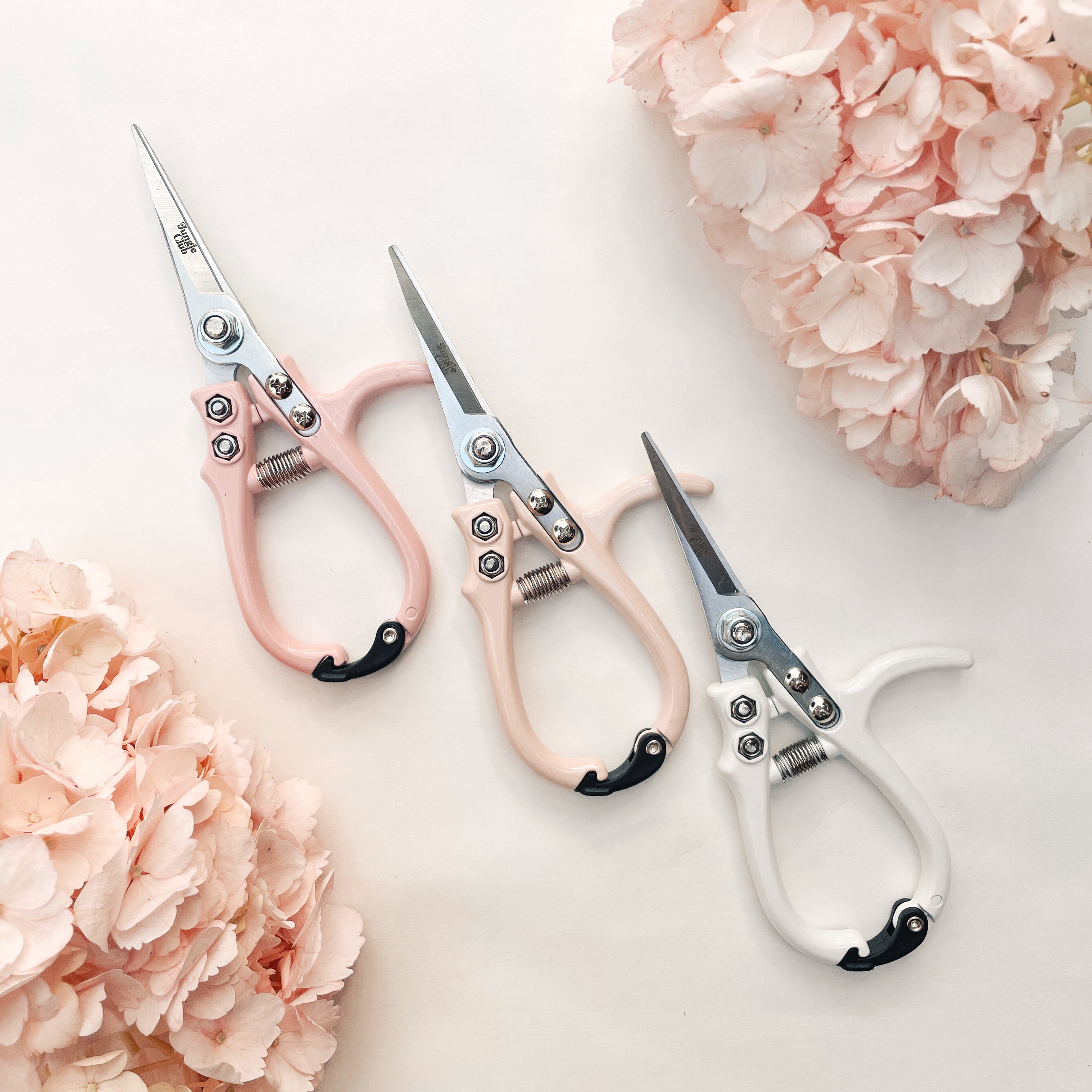 All three color ways of the pruning shears photographed next to each other and staged in the middle of hydrangeas. There is a white, a light pink and a neutral tan. 