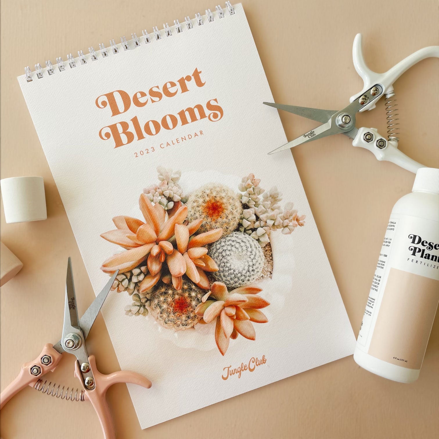A spiral bound calendar with a cream front cover with a succulent and cacti arrangement photograph and &quot;Desert Blooms 2023 Calendar&quot; in burnt orange text on the top. The interior features a monthly calendar layout accompanied by a different succulent or cacti photograph on each month&#39;s page. 