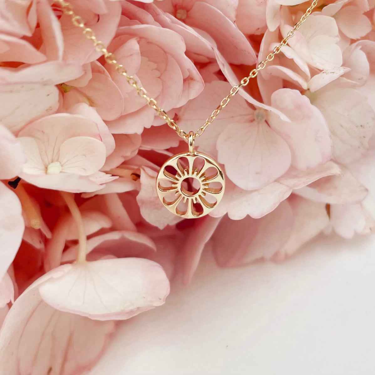 A dainty gold chain necklace with a gold circle pendant in the center with a cut out of a daisy flower, photographed in front of pink hydrangeas.