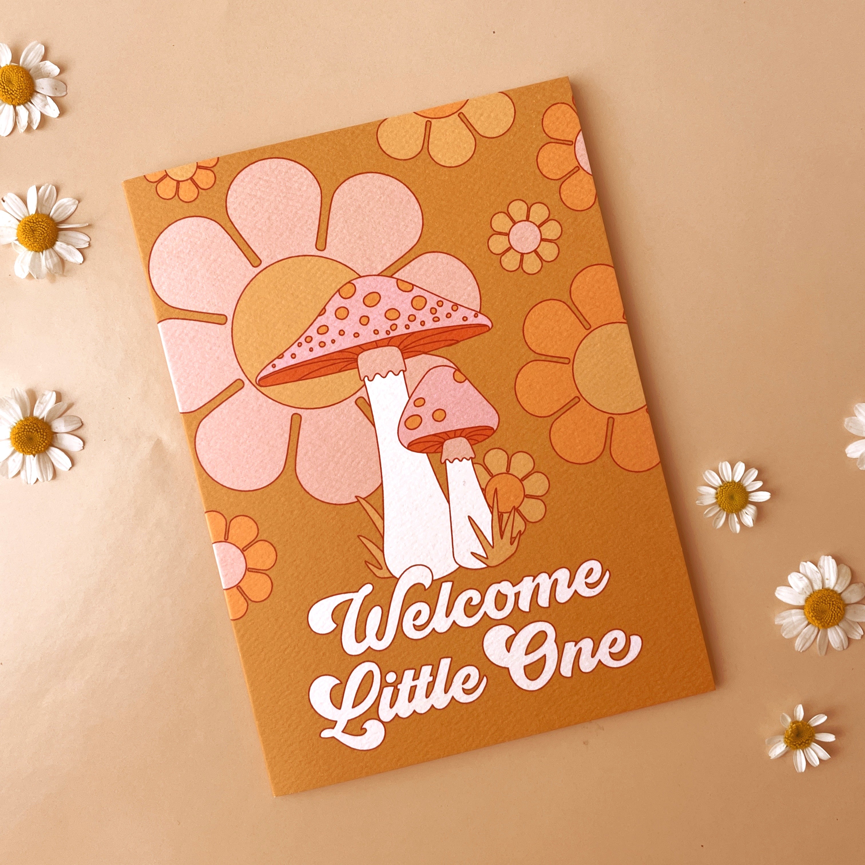 On a cream background is an orange card with a floral and mushroom design along with white text that reads, &quot;Welcome Little One&quot;.