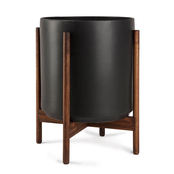 This cylinder pot is solid black and sits within four spokes of a dark walnut wood plant stand, standing about 5.5 inches from the ground.