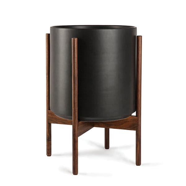 This 5 gallon, cylinder pot is solid black and sits within four spokes of a dark walnut wood plant stand, standing about 7 inches from the ground.