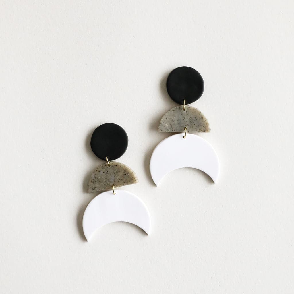 Photo of a pair of earrings featuring a white moon hanging from a grey speckled half circle and a black circle on top. Made out of clay. Earrings are on a white background.