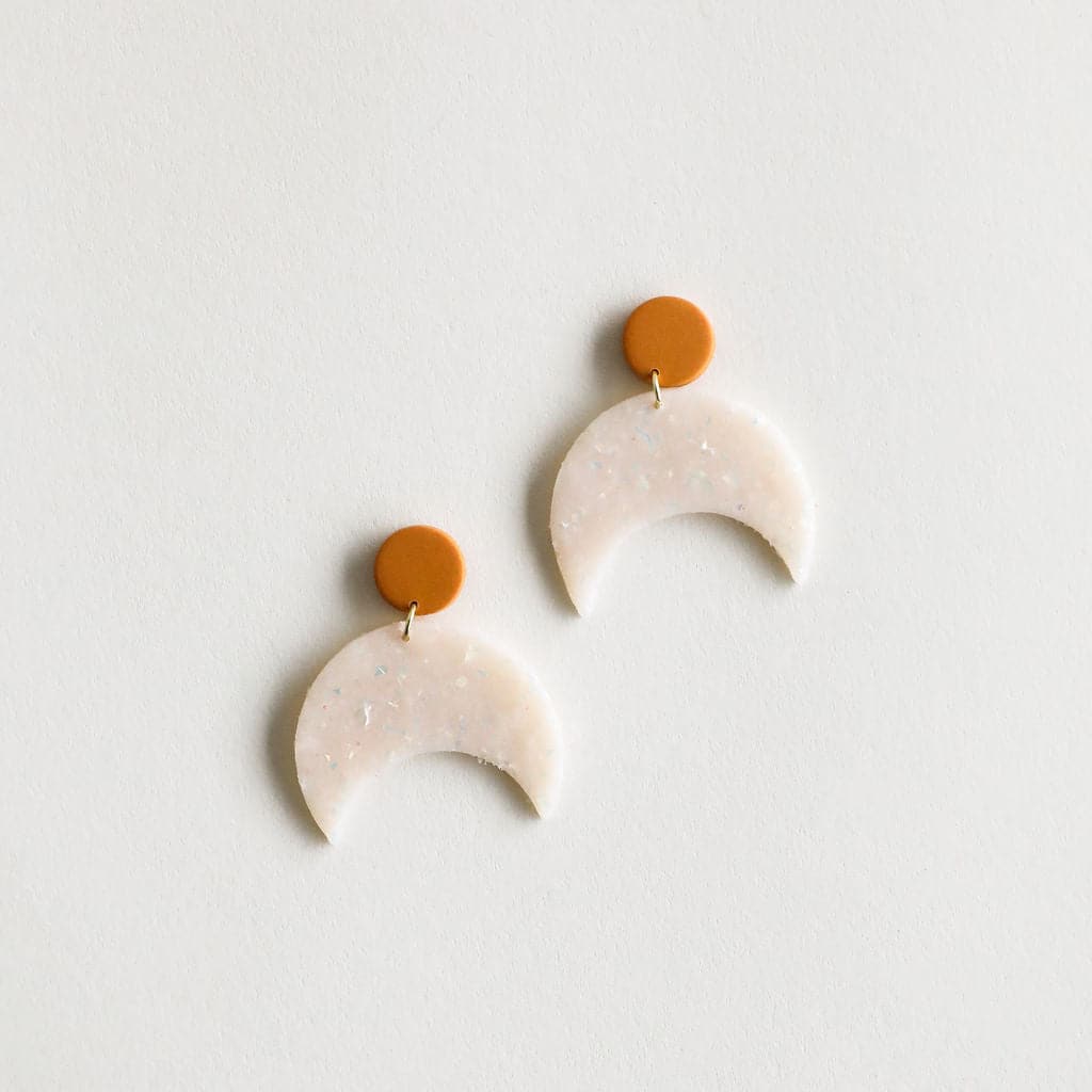 Earrings with a opalescent moon hanging off of a small orange circle on a white background.