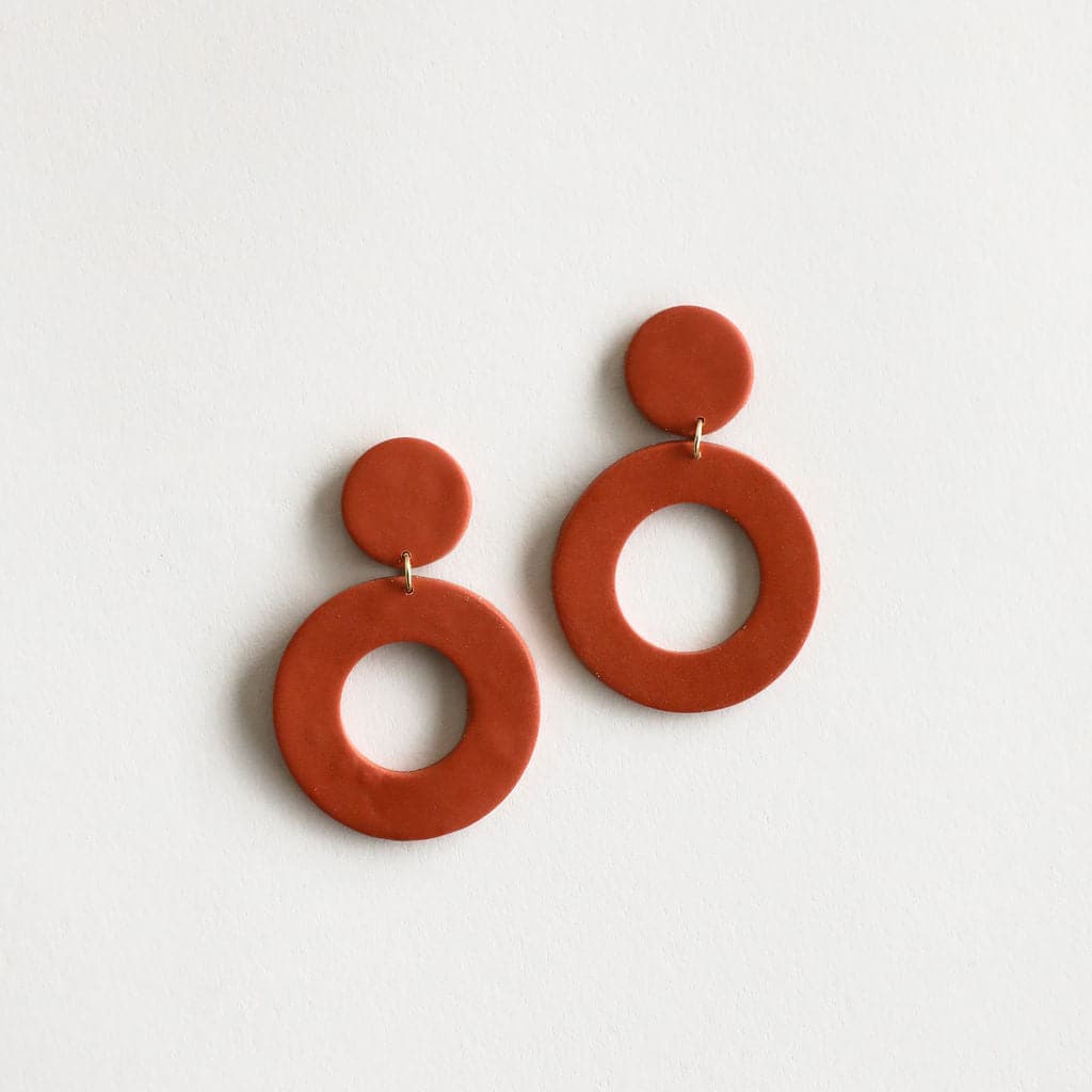 The Mollye earrings that feature a duel circle design. The top circle is smaller and solid while the lower circle is larger and open in the center. This earring features a straight post backing and comes in a variety of colors. This color way is solid rust red.