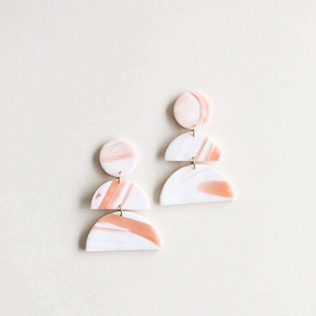 In front of a white background is a pair of earrings. The top is a white and light rust marble circle. Bellow is a half circle of the same color and below that is a larger half circle of the same color. Each piece is connected with little gold rings.