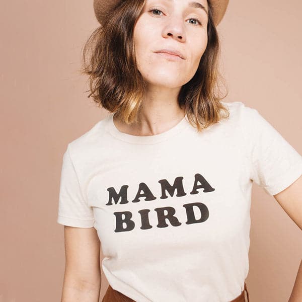 A cream cotton t-shirt with black letters across the from that read, &quot;Mama Bird&quot; worn on a model in front of a tan background.