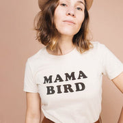 A cream cotton t-shirt with black letters across the from that read, "Mama Bird" worn on a model in front of a tan background.