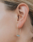 a close up of a woman wearing gold bent wire drop earrings with ammonite gemstone bar.
