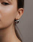 Medium sized silver hoops that have a wider frame and a thin depth. They feature a straight post and a clasp on the back top of the hoop to secure it on your ear.