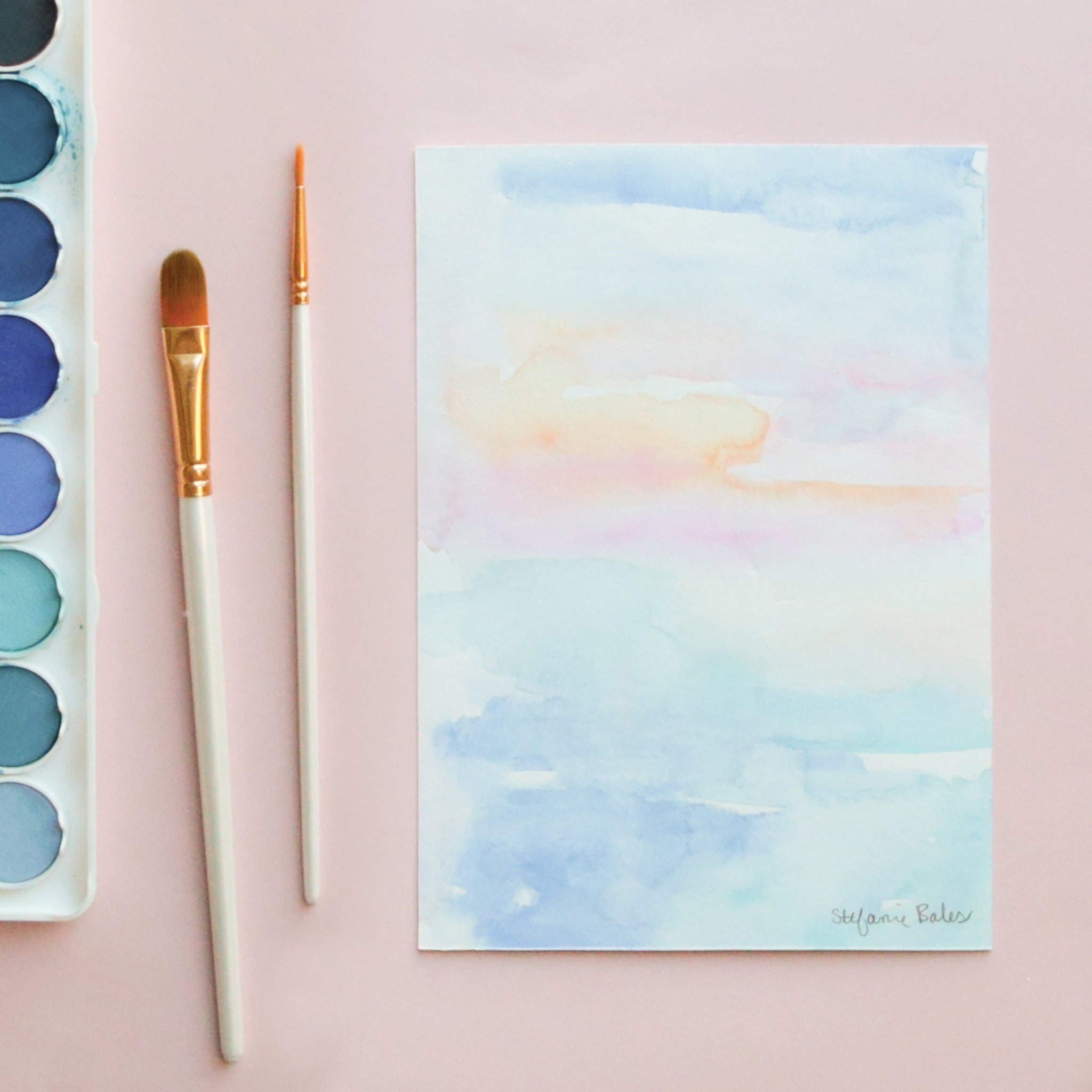 On a light pink background is a photo of blue toned watercolors and paintbrush alongside a piece of art next to it.