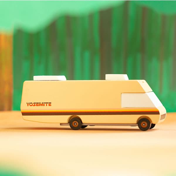 Miniature RV beige beech wood with white windows and dark brown wheel detailing. An orange and brown vintage stripe accent runs across its side. The RV models lays against a blurred emerald green forest scene.  