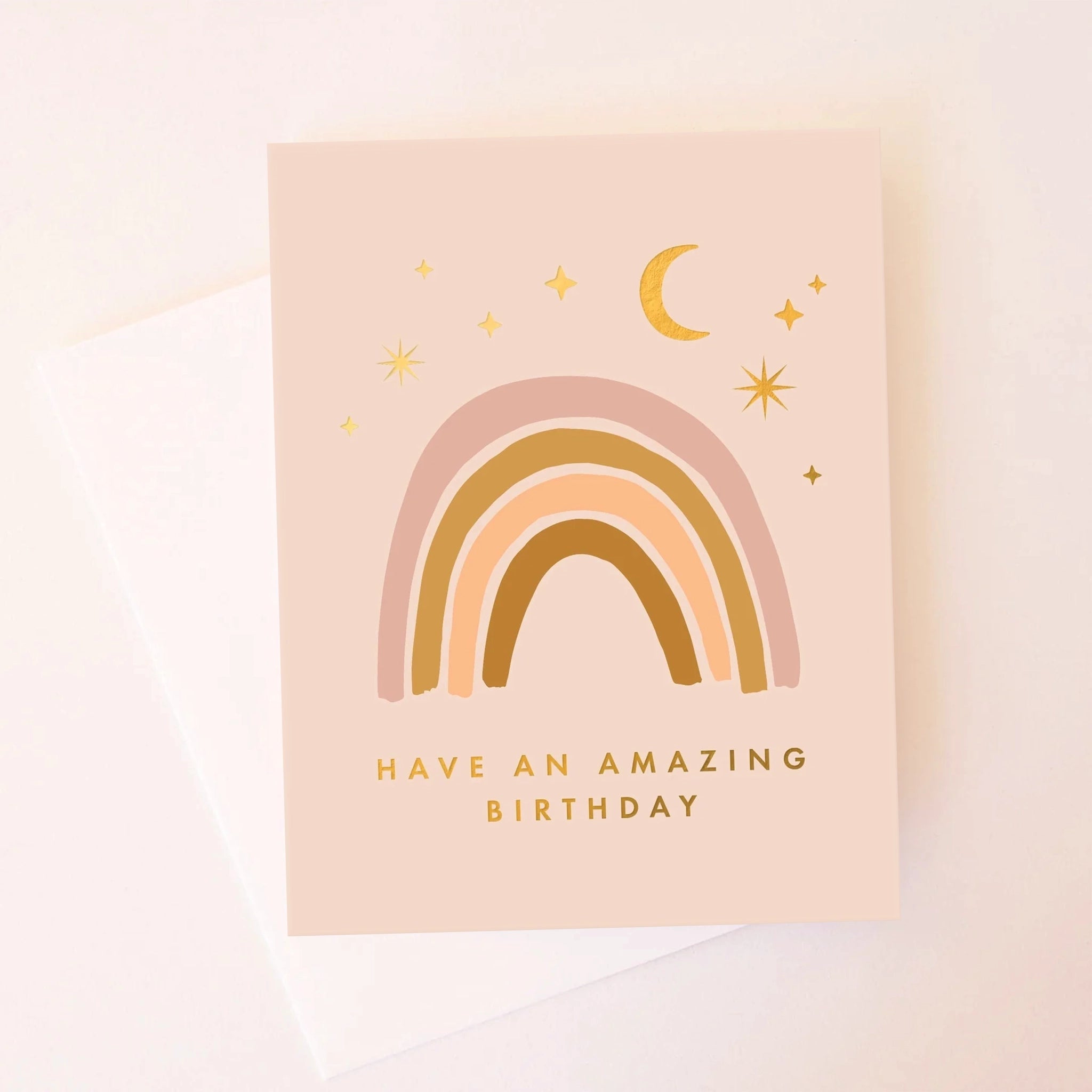 A light pink card with a neutral rainbow on the front under moon and star designs and gold text along the bottom that reads, "Have an amazing birthday" as well as a coordinating white envelope.