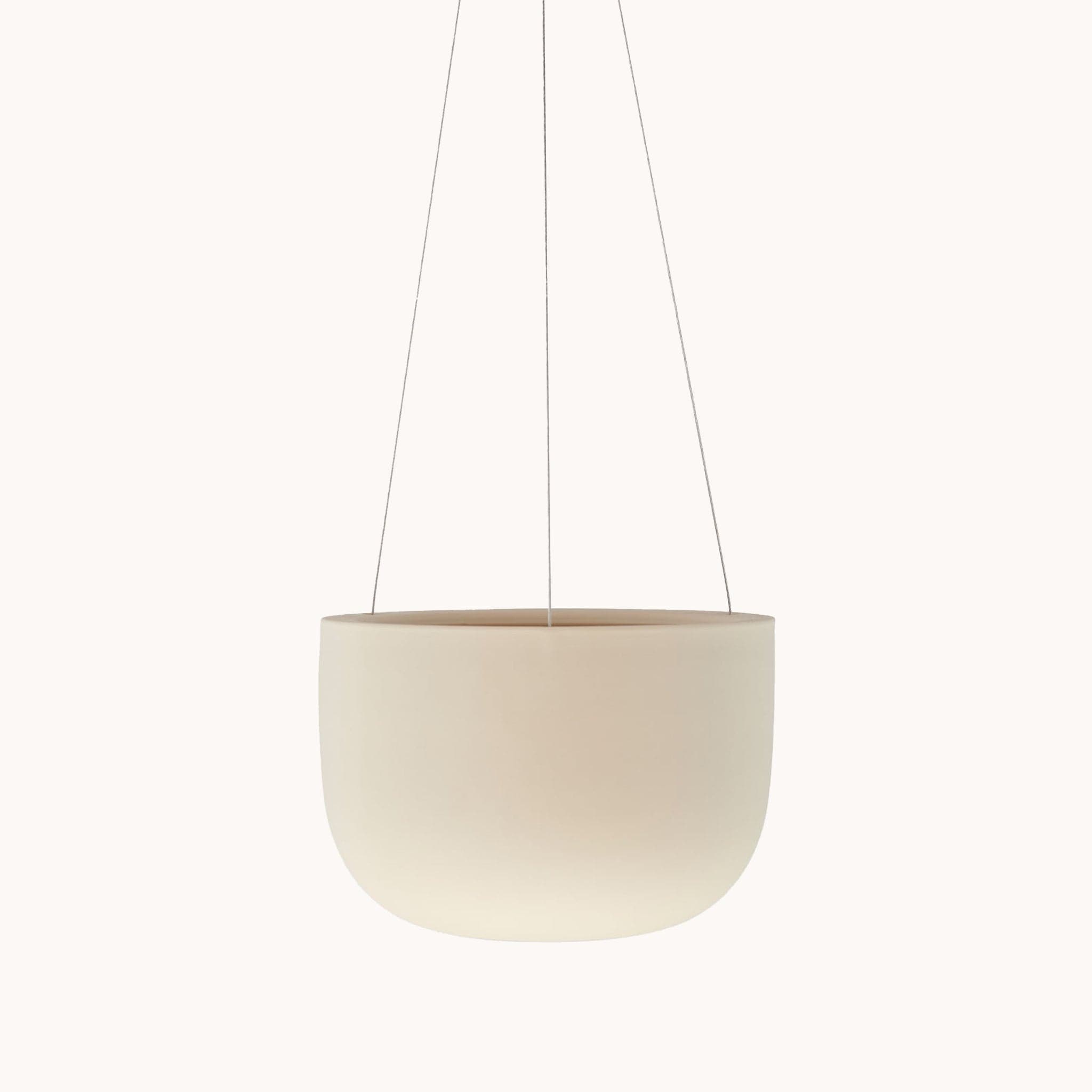 Hanging in front of a white background is a light tan hanging pot. The pot is wide and round and slightly tapers at the bottom. The pot is being held by three silver wires.