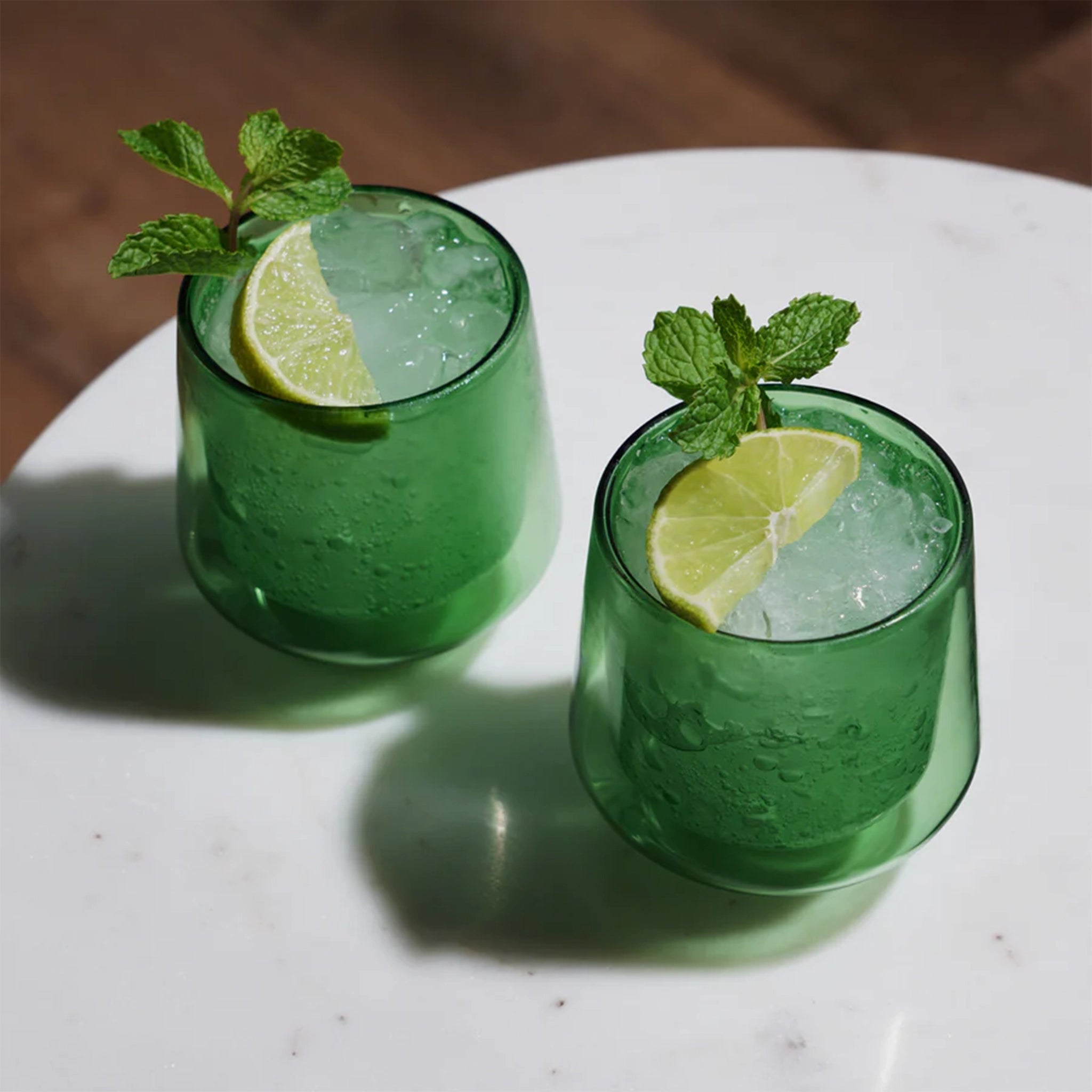 A set of two double walled glass tumbler cups in a green color photographed with a drink inside and garnished with lime and mint. 