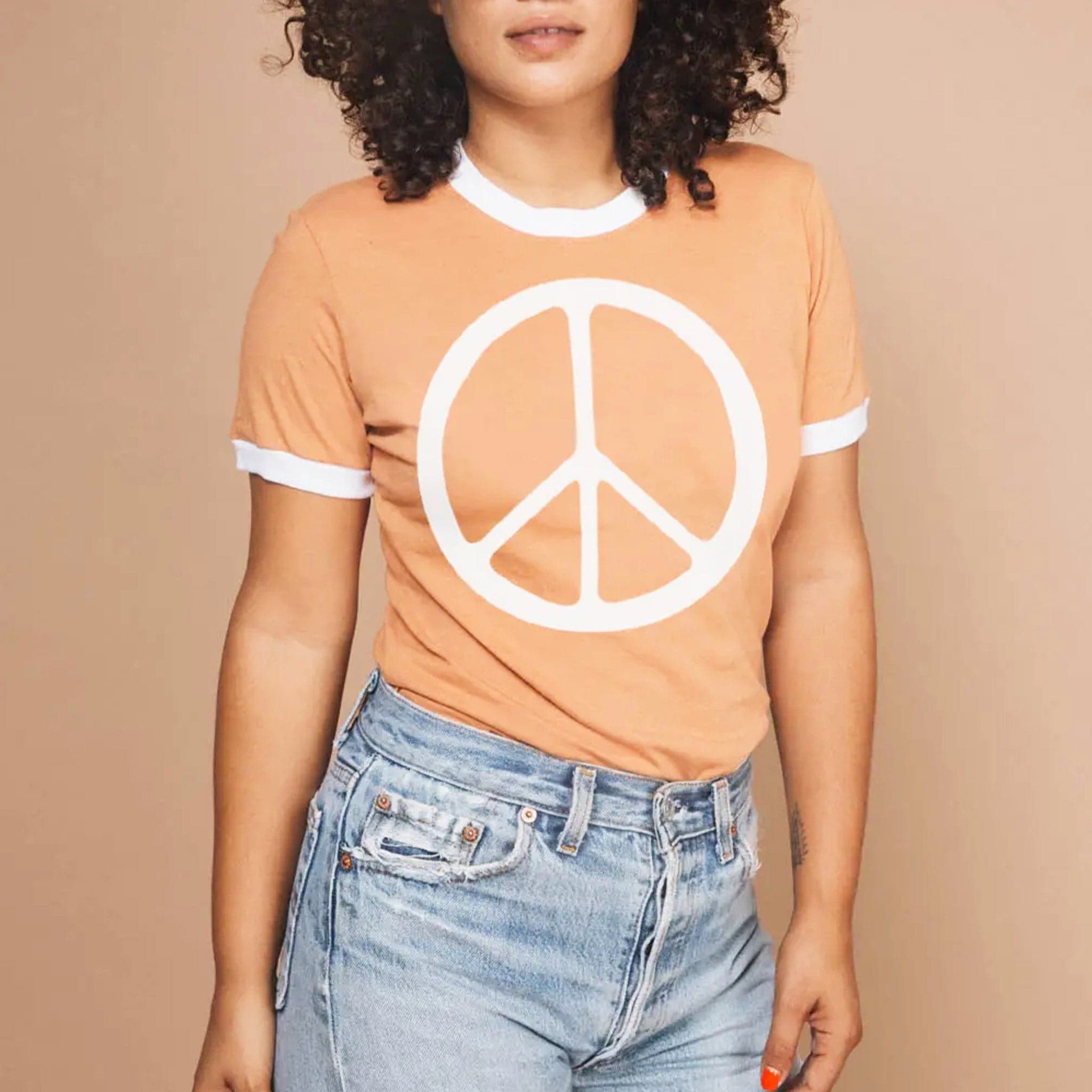 In front of a peachy background is a woman wearing a light orange short sleeve t-shirt. The collar and end of the sleeves have a white border. In the middle of the shirt is a large white peace sign. 