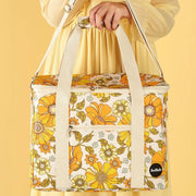 Photographed in front of a yellow background is a picnic bag with cream straps and 70's style yellow and orange floral print. 