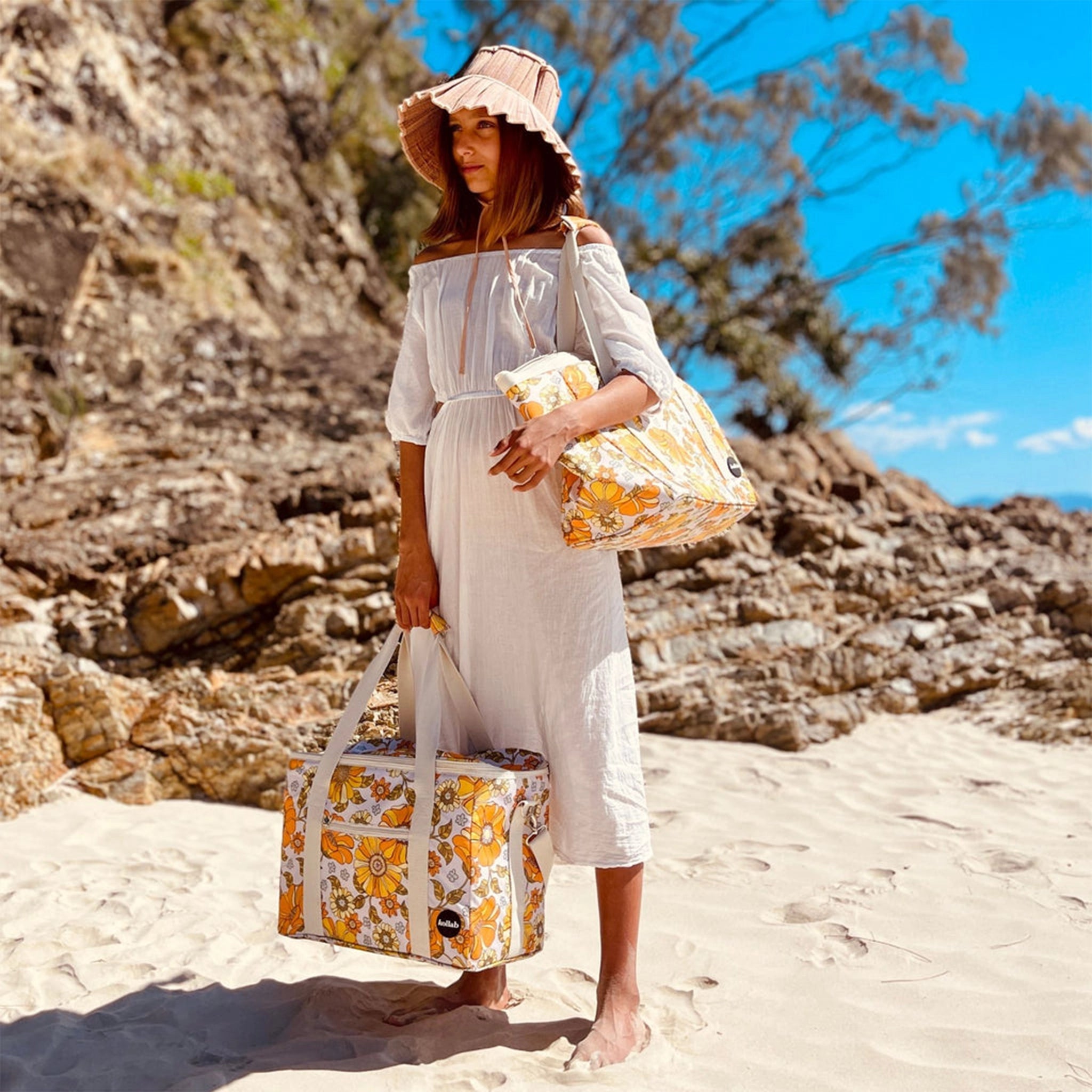 Photographed in front of a yellow background is a picnic bag with cream straps and 70's style yellow and orange floral print held by a model on a beach.