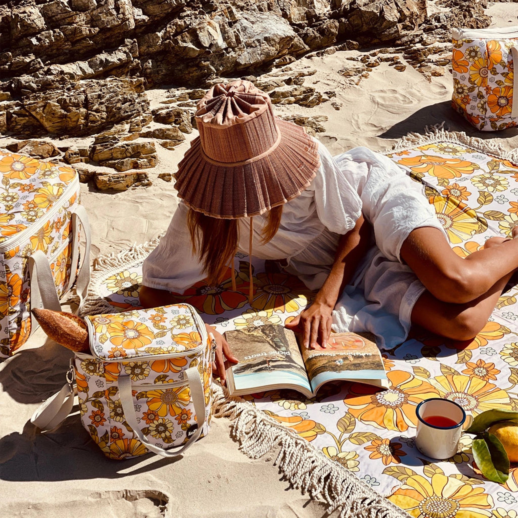 A 70&#39;s style yellow and orange floral picnic mat with cream fringe detailing around the edges photographed with coolers and beach bags in the same print with a model laying on it reading a book.
