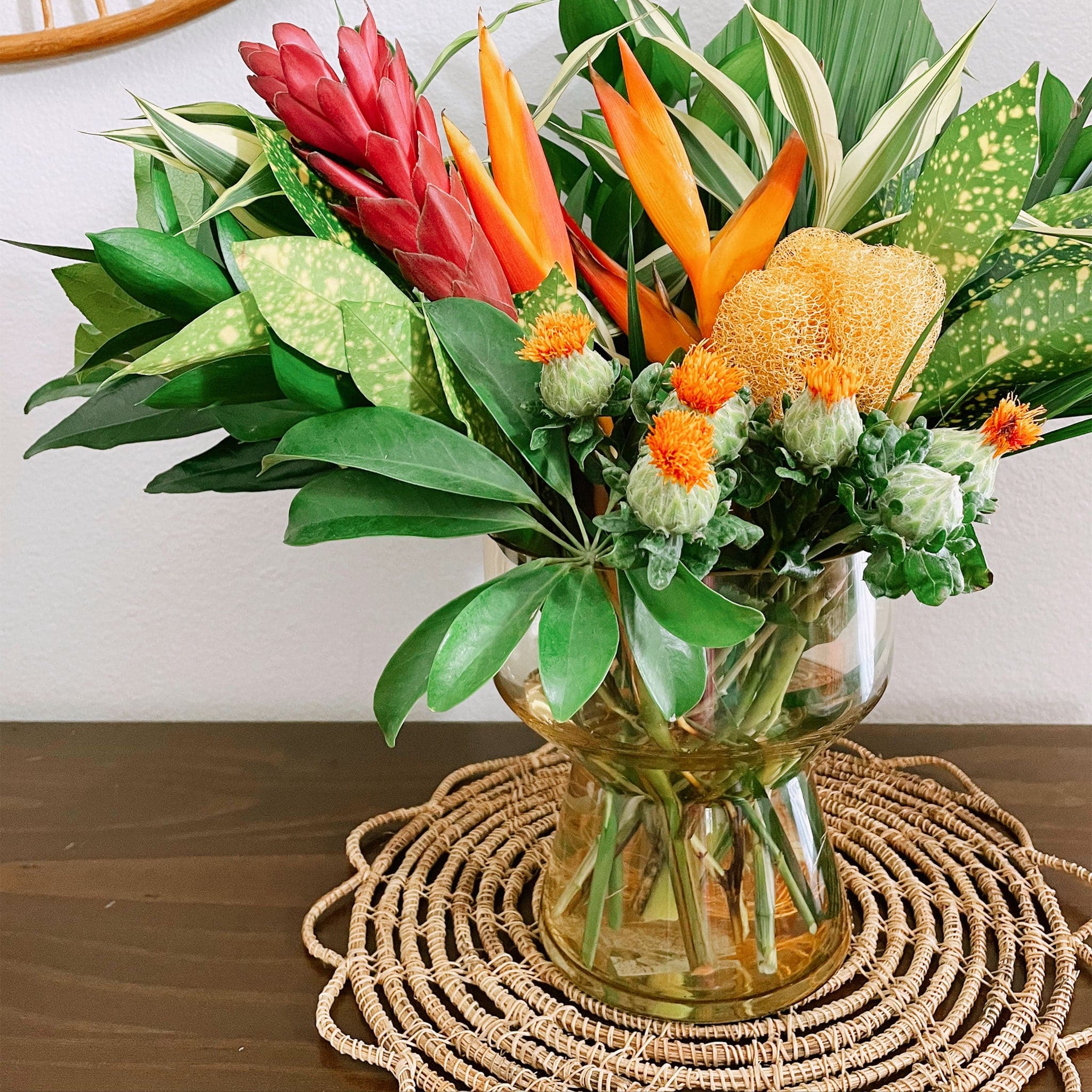 An amber colored compote style glass vase filled with a bouquet of tropical flowers on a woven placemat on a wood tabletop.