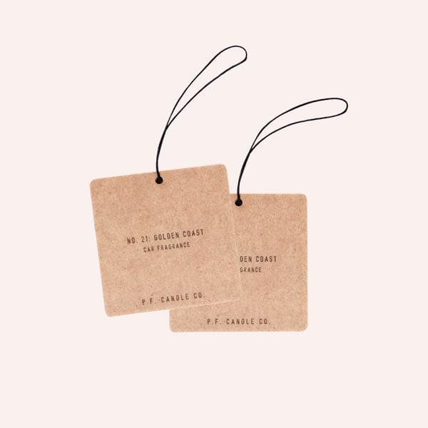 Two cardboard square air fresheners with a black elastic loop for hanging and small black text that reads, &quot;No. 21: Golden Coast Air Freshener, P.F. Candle Co.&quot;.