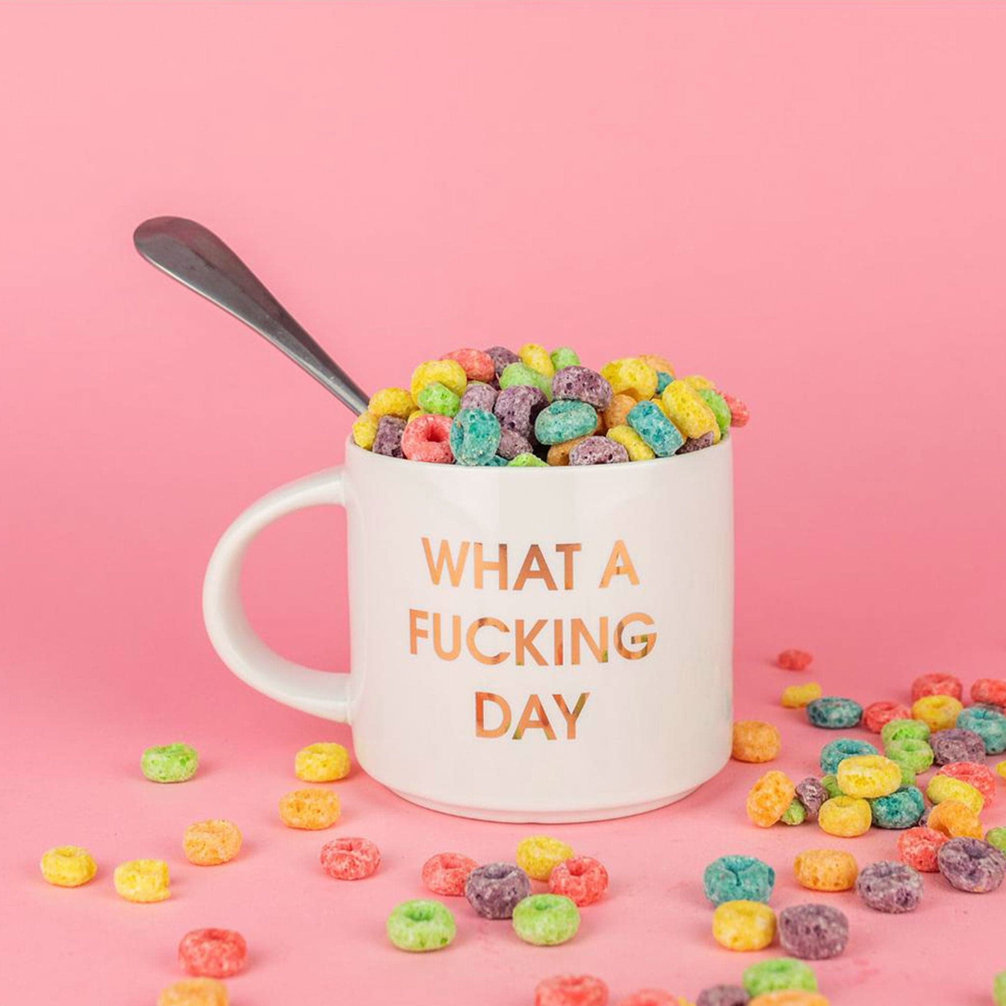 Classic white ceramic mug labeled &#39;What a Fucking Day&#39; in gold shimmering capital lettering. Mug has a round white handle fasted on the left hand side. Mug is filled with overflowing, rainbow colored fruit loops. Behind lays a bright pink background.
