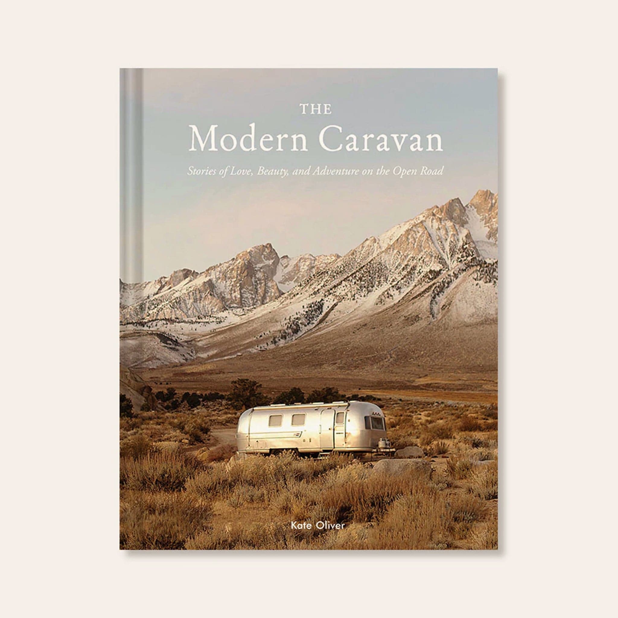 Hard cover of travel book titled &#39;The Modern Caravan, stories of love, beauty, and adventure on the open road&#39; in white pressed lettering. Behind the title is a cool toned mountain scene. Below the mountains sits a silver airstream trailer amidst an open valley.
