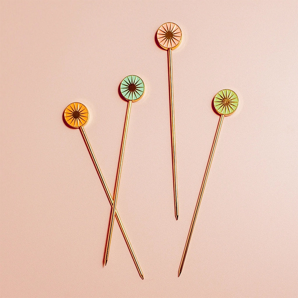 Four cocktail picks with a round top with a sun design in four different shades of green, teal, salmon pink and orange.