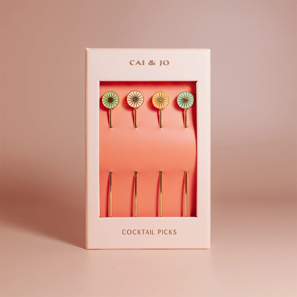 Four cocktail picks with a round top with a sun design in four different shades of green, teal, salmon pink and orange. This photograph shows them packaged in their box with a pink border. 