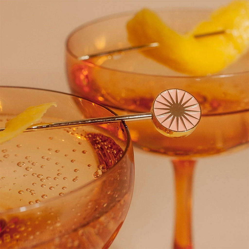 Four cocktail picks with a round top with a sun design in four different shades of green, teal, salmon pink and orange. This photograph shows the pick holding up an orange slice garnish above the beverage.