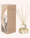 A oil diffuser with a clear glass filled with scented oil a clean cream colored lid and five neutral wood reeds stemming out from the top next to the decorative box that the diffuser comes in which is a tan box with floral designs on the sides.