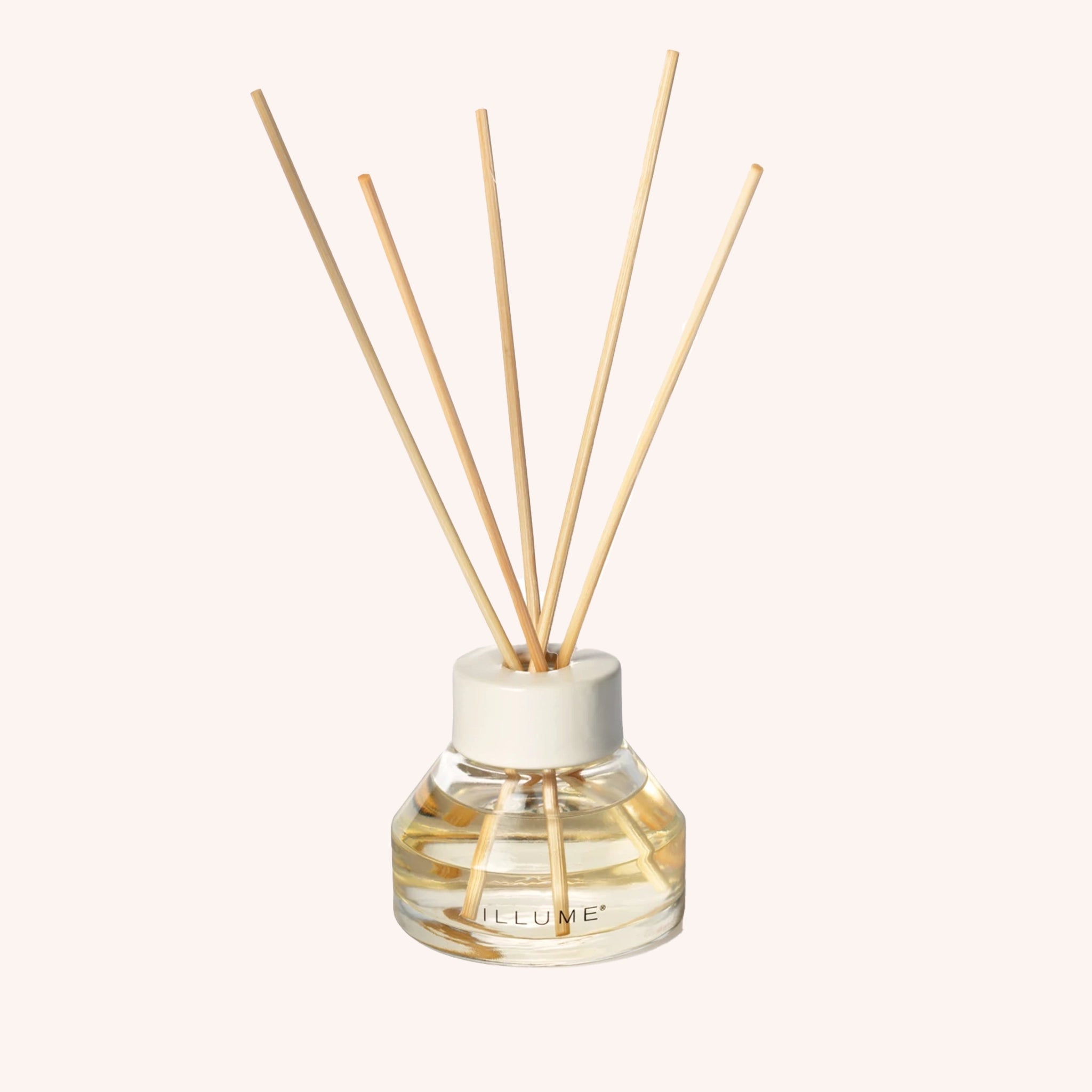 A oil diffuser with a clear glass filled with scented oil a clean cream colored lid and five neutral wood reeds stemming out from the top.