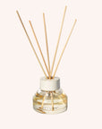 A oil diffuser with a clear glass filled with scented oil a clean cream colored lid and five neutral wood reeds stemming out from the top.