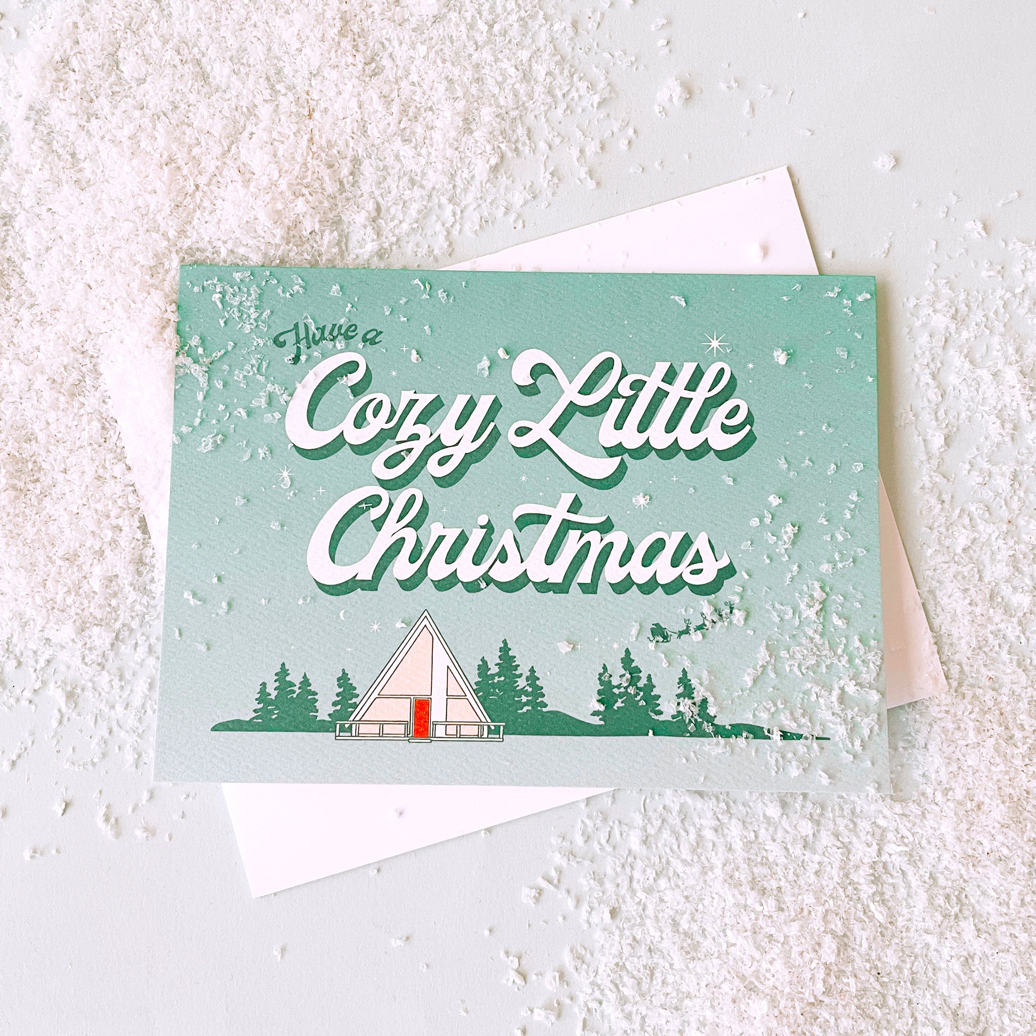 Aqua and white ombre greeting card that reads 'have a cozy little Christmas'. Below the text is a small white tent in a forest scene. A small Santa sleigh and reindeer fly across the sky. The card is accompanied by a white envelope.